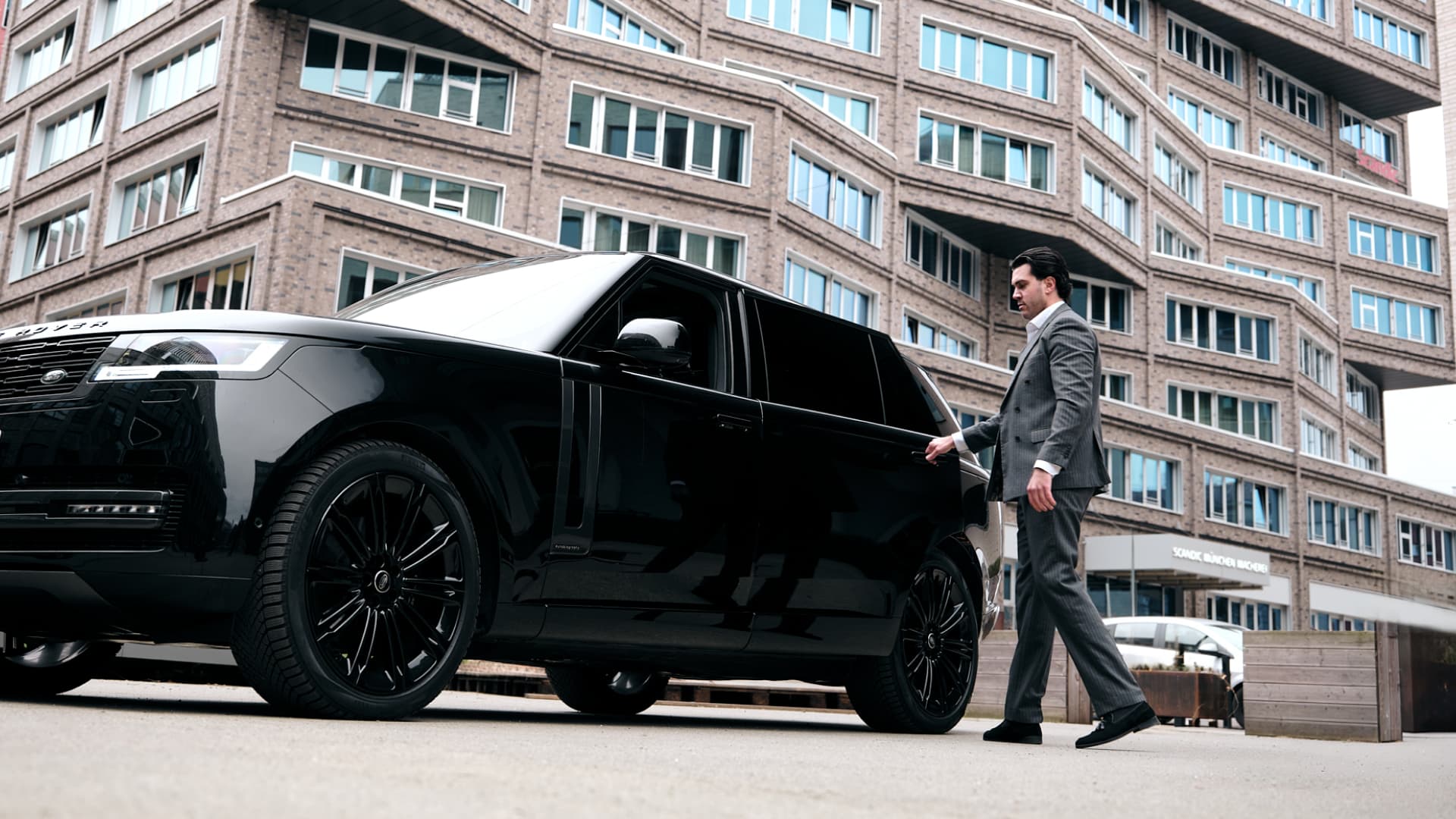 Chauffeur in a suit opening the door of a Range Rover in front of the Scandic Hotel at Macherei Munich