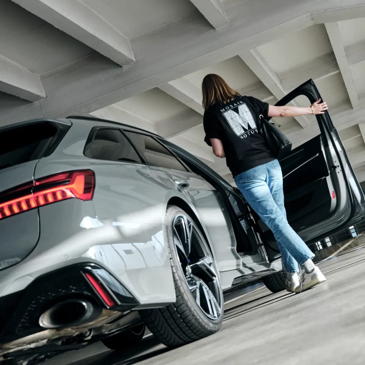 Close-up of a grey Audi RS6 in a parking garage next to a woman wearing a Mosaik Motors branded shirt.
