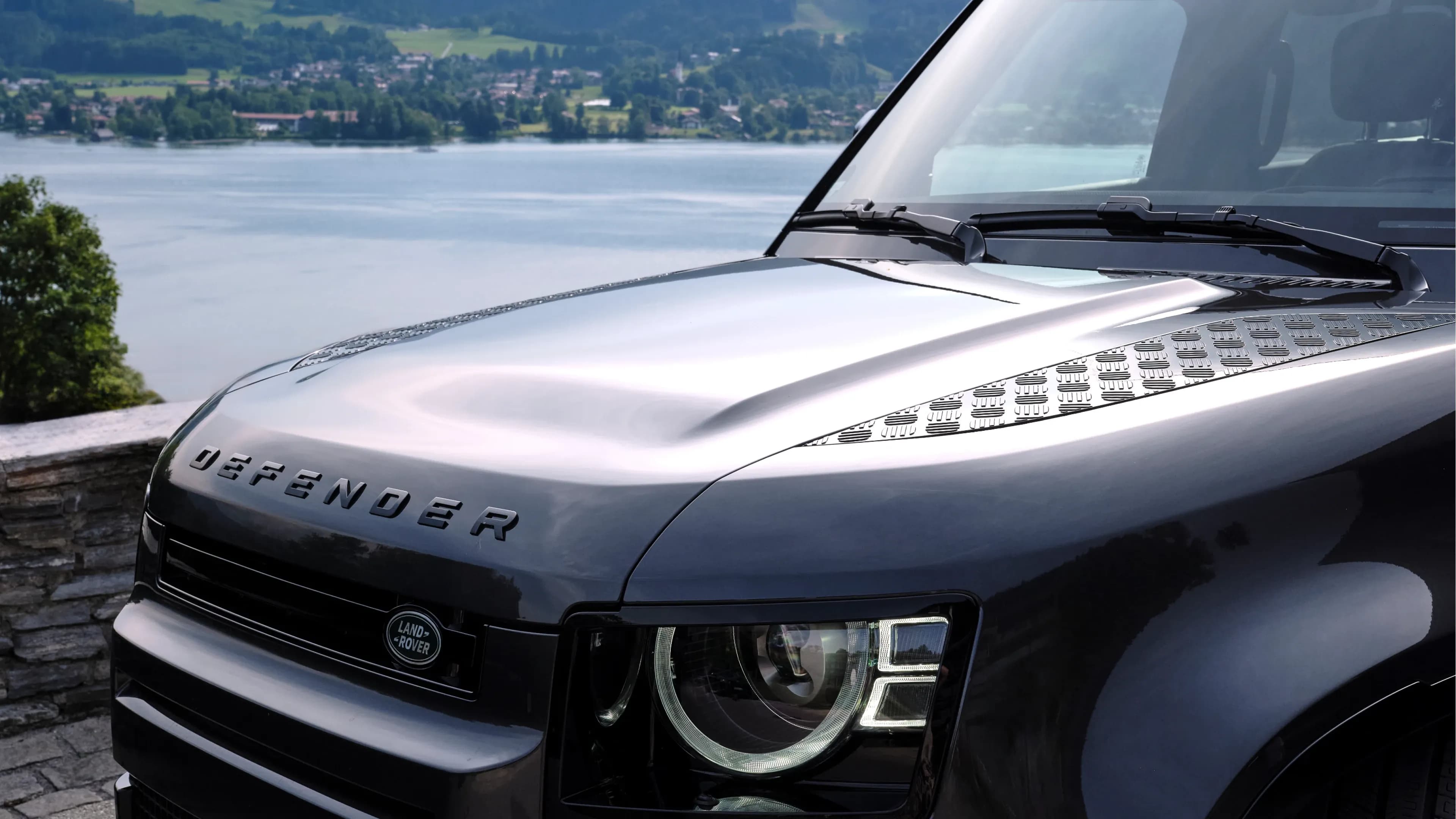 Grauer Land Rover Defender 110 von Mosaik Motors mit Fokus auf der Motorhaube, im Hintergrund der Tegernsee und die bayerischen Alpen.