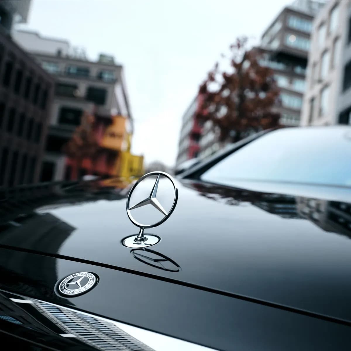 Rent a Mercedes-Benz S-Class in Munich – close-up of the iconic Mercedes hood star with the modern Macherei design offices in the background by Mosaik Motors.
